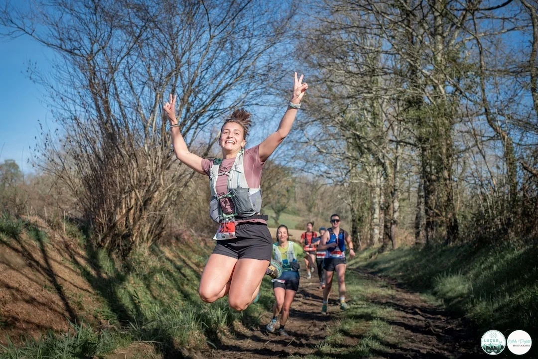Femme en plein saut, souriante, lors d’un trail nature sur les sentiers de Lucq-de-Béarn, entourée d’autres coureurs.